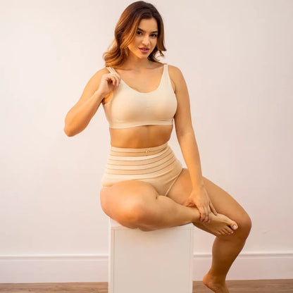 Imagem de uma mulher sentada, usando um conjunto de sostén y calzón moldeador Model - Seca Barriga, ambos na color beige. A calzón possui listras horizontais y uma flor delicada na faixa superior. Ela sorri y segura a tirante do sostén enquanto está em uma pose descontraída sobre um cubo blanco.