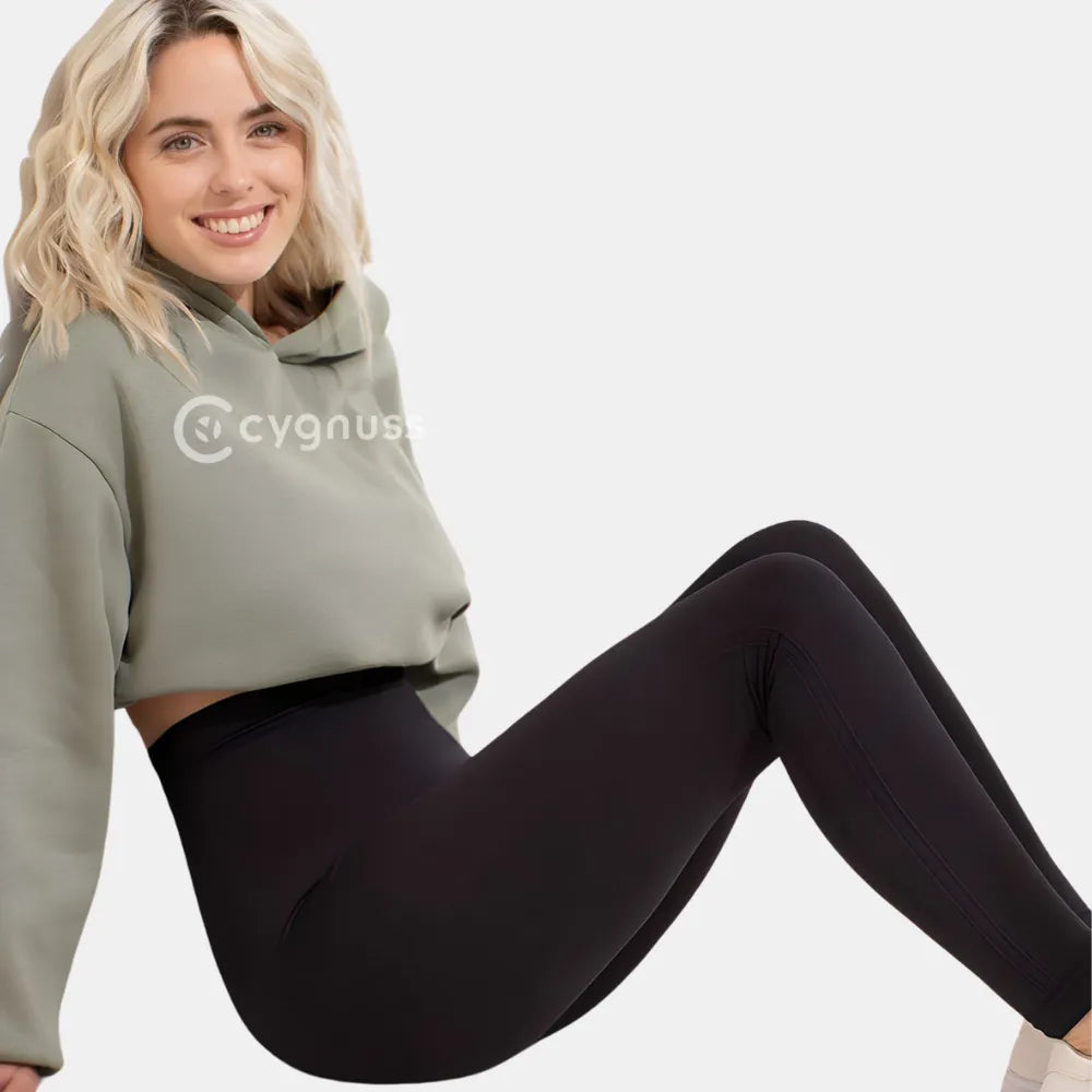 Mulher sorrindo usando a Legging Jade Fit moldeador de Cós Alto na color preta, combinada con um hoodie verde, sentada de forma descontraída, destacando o cós alto y o ajuste cómodo da peça.