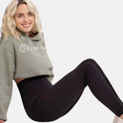 Mulher sorrindo usando a Legging Jade Fit moldeador de Cós Alto na color preta, combinada con um hoodie verde, sentada de forma descontraída, destacando o cós alto y o ajuste cómodo da peça.