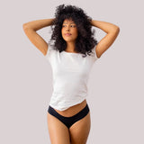 Mulher de cabelos cacheados posando con as mãos no cabelo, vestindo uma camiseta branca y uma calzón fio-dental preta sin costuras da Cygnuss. A imagem destaca o ajuste cómodo y a elegância minimalista da peça