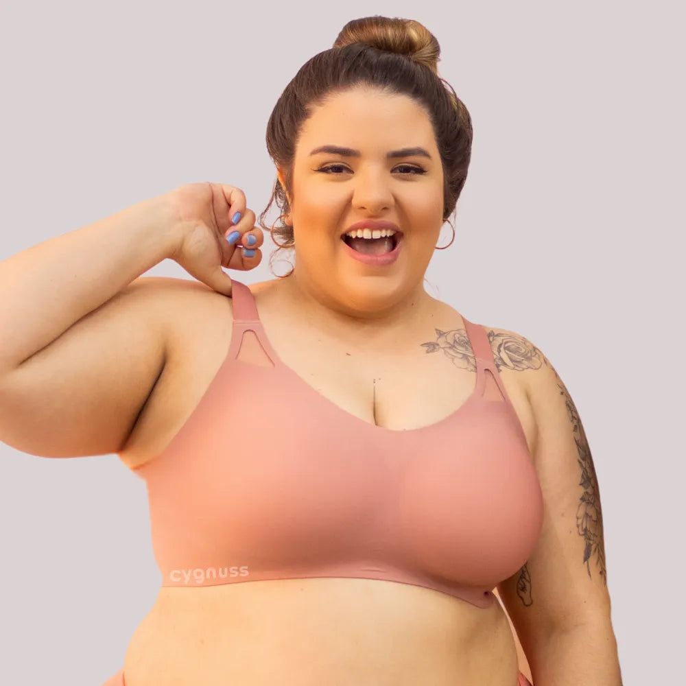 Mulher plus size sorridente vestindo o sostén Dreams na color rosé, destacando o design sin costuras, as tirantes largas y os detalhes vazados próximos aos hombros. O tejidos molda o corpo con comodidad y sustentação. O logo da Cygnuss está visível na parte inferior do sostén.